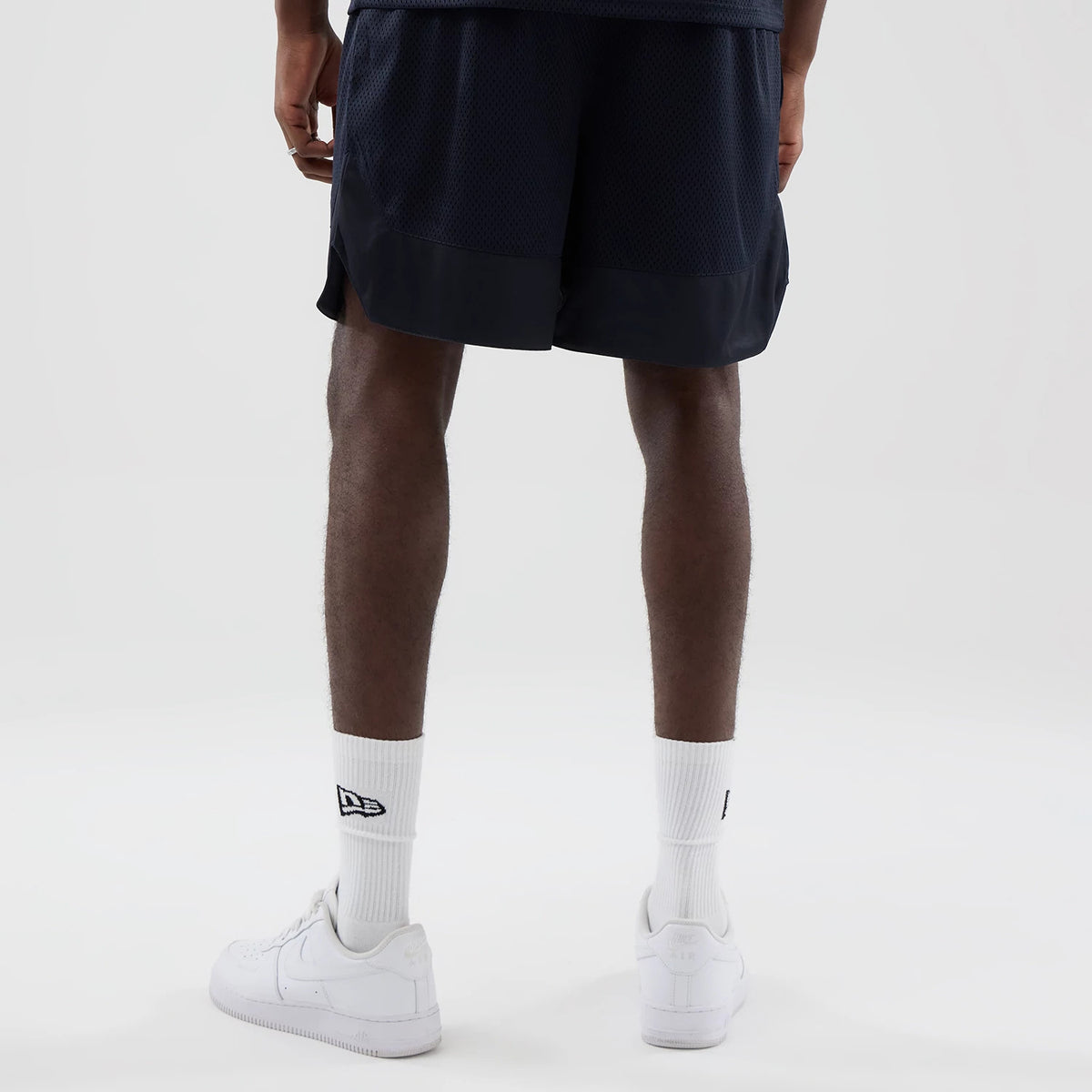 New Era Mesh Navy Oversized Shorts, back view on a model showing the mesh texture with a solid navy band at the hem, paired with white trainers and socks featuring a black embroidered New Era logo on the side.
