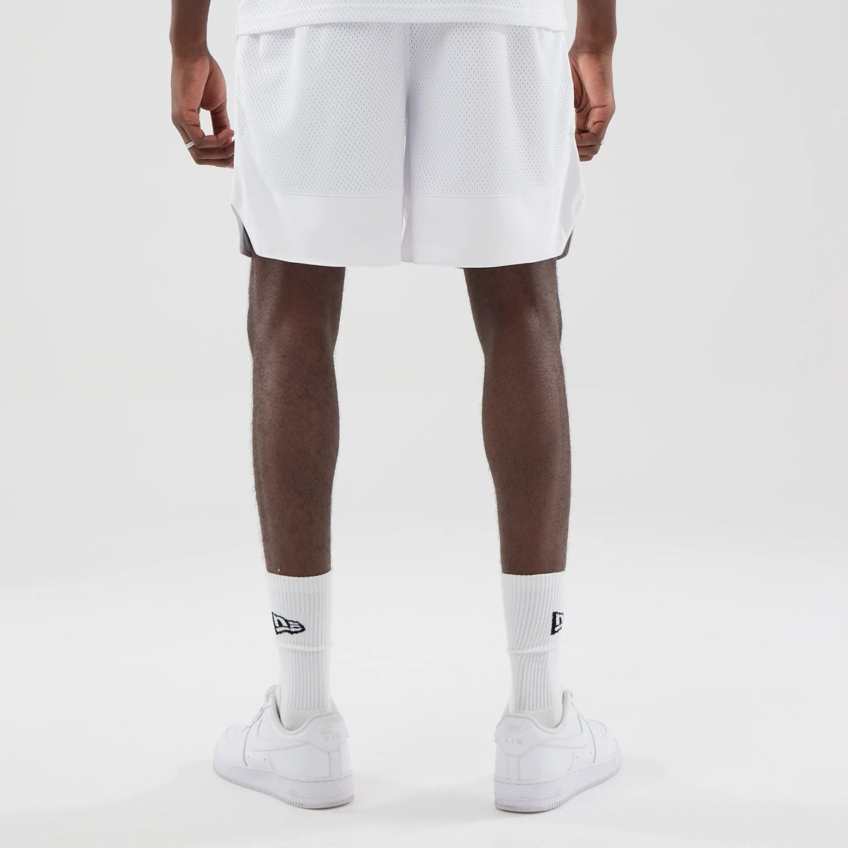 New Era Mesh White Oversized Shorts, back view on a model showing the white mesh fabric with a solid white hem, loose fit design, and side splits at the hem, styled with white socks featuring an embroidered New Era logo and white trainers.