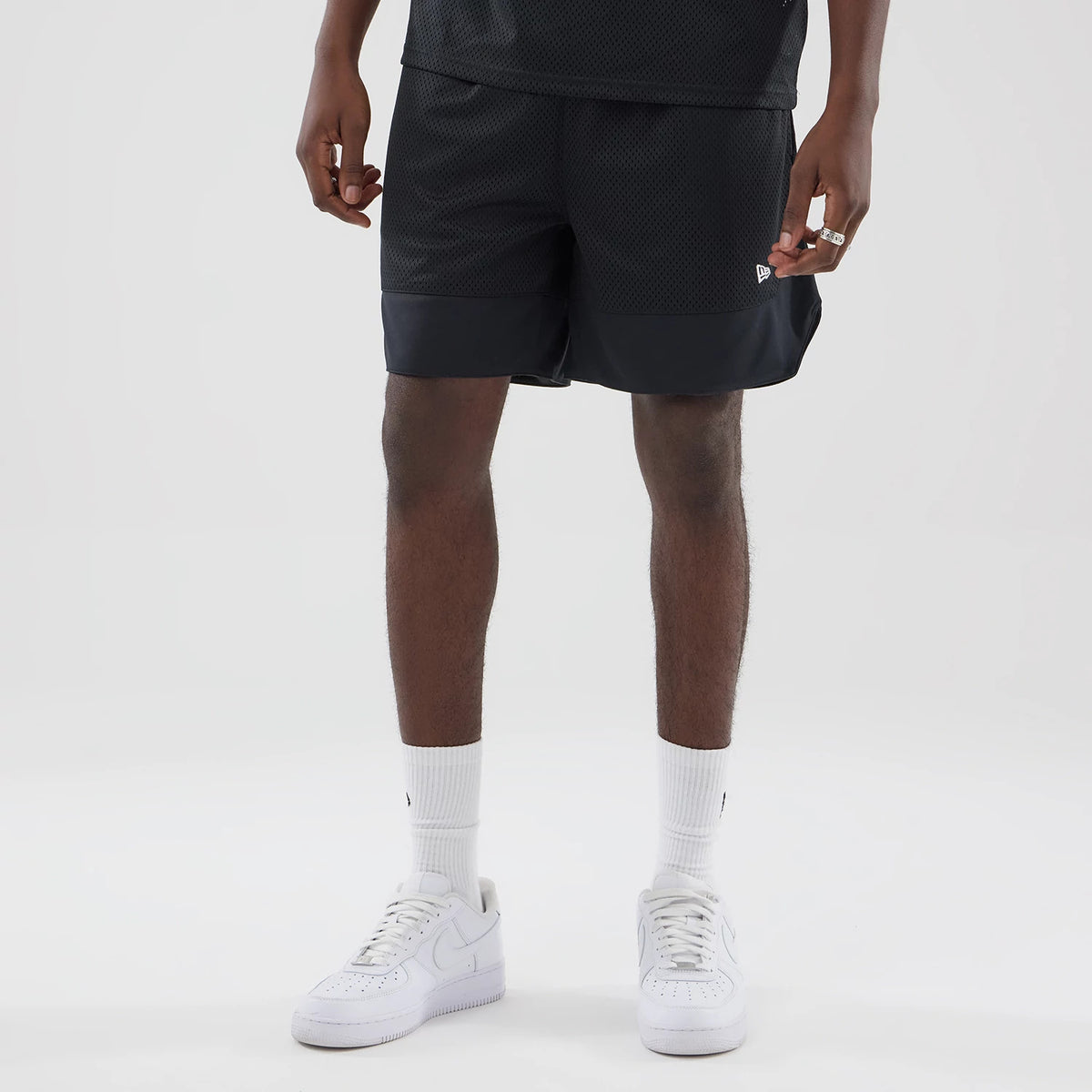 New Era Mesh Stripe Black Oversized Shorts, front view on-model showing a mesh texture with a contrasting black band at the hem, loose fit above the knee, and a small white embroidered New Era logo near the left hem.