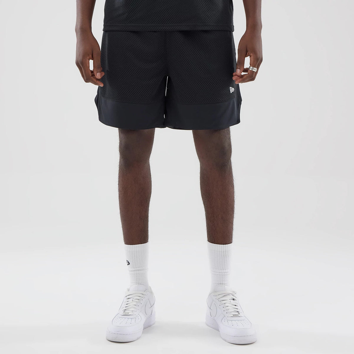 New Era Mesh Stripe Black Oversized Shorts, front view on a model, showing the black mesh fabric with a stripe across the bottom, an embroidered New Era logo on the left leg, and styled with white socks and trainers.