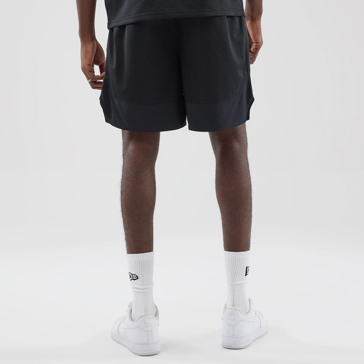New Era Mesh Stripe Black Oversized Shorts, back view on a model showing the mesh texture and contrasting solid black panel at the hem, with the model wearing white socks featuring the New Era embroidered logo and white trainers.