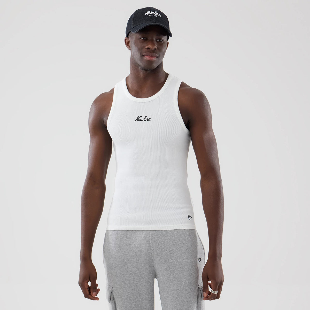 New Era Rib White Vest, front view on a model wearing a black cap, showing the black New Era script logo centred on the chest, ribbed texture of the fabric, and a small New Era flag logo at the lower left hem.