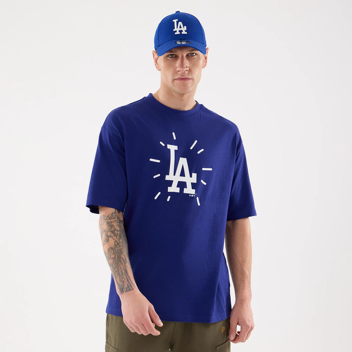 New Era LA Dodgers MLB 59FIFTY Day Graphic Dark Blue T-Shirt, front view on a model styled with matching blue cap, featuring a large white printed LA logo at the centre of the chest with surrounding lines radiating outward.
