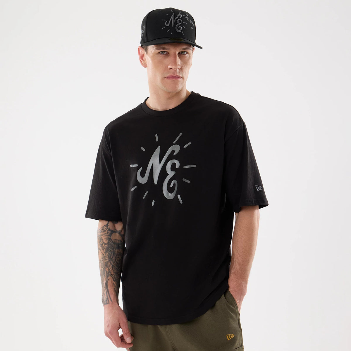 New Era 59FIFTY Day Graphic Black T-Shirt, front view on a model paired with a black cap, showing a large silver graphic NE on the chest and an embroidered New Era logo on the left sleeve near the hem.