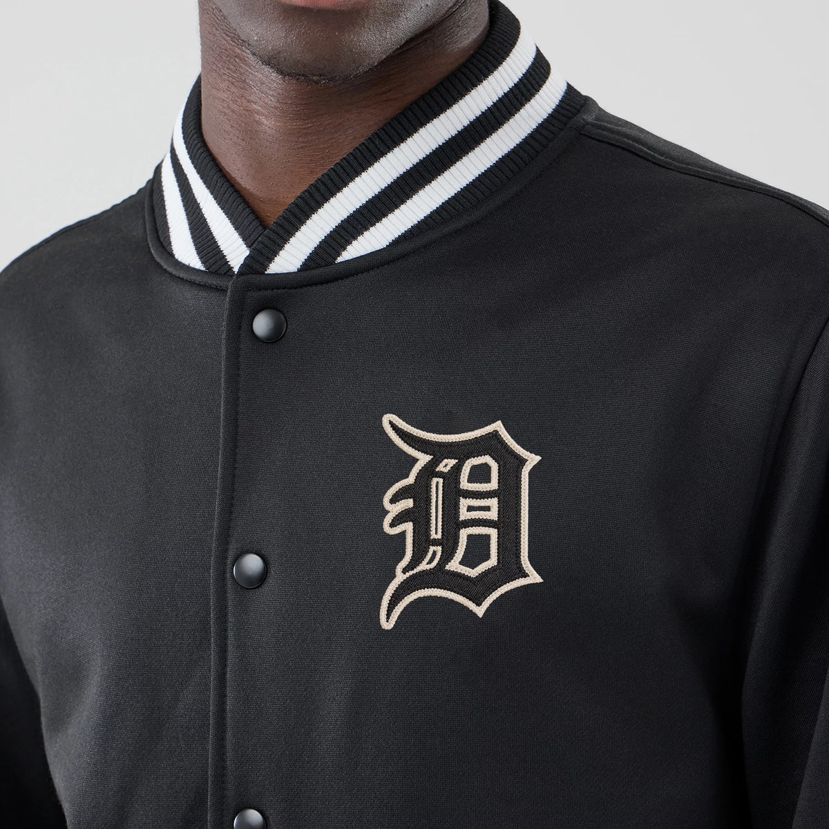 New Era Detroit Tigers MLB Jersey Bomber Black Jacket, front view showing the cream embroidered Detroit Tigers logo on the left chest, black snap buttons down the front, and a ribbed black and white striped collar on a model.
