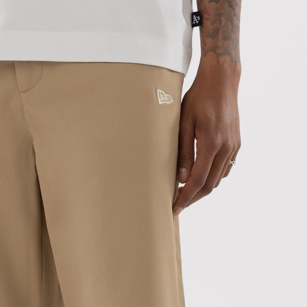 New Era Athletics MLB Beige Woven Chinos, close-up on a model showing the beige fabric with a texture, the New Era flag logo embroidered on the left thigh, and a white T-shirt partially at the top of the image.
