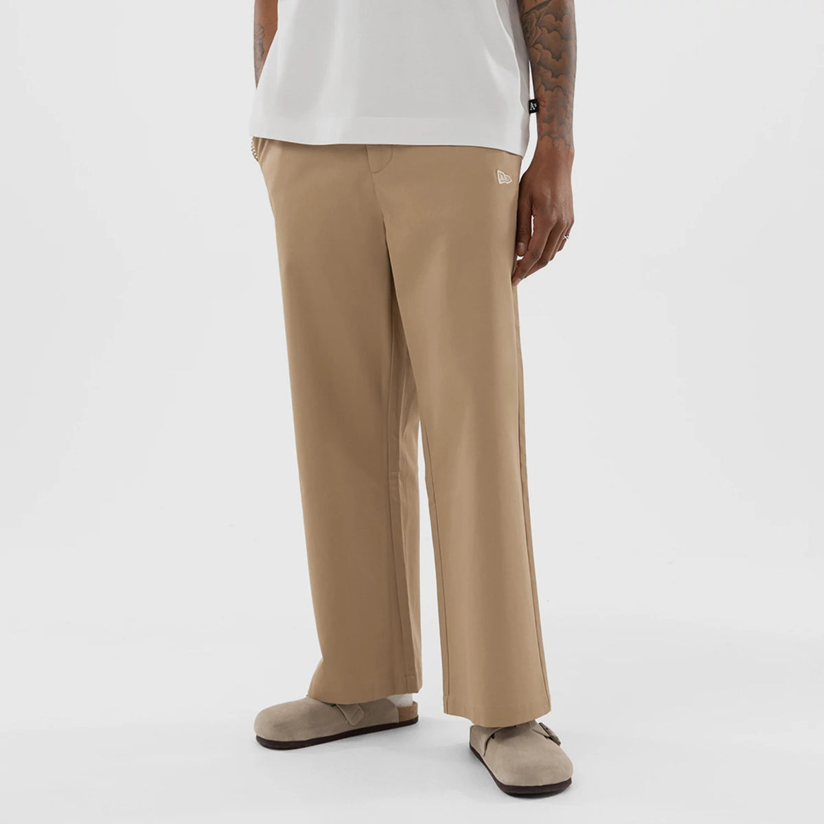 New Era Athletics MLB Beige Woven Chinos, front view on a model showing the relaxed fit with straight legs, the New Era flag logo embroidered on the left hip, and the beige colour paired with neutral slip-on footwear.