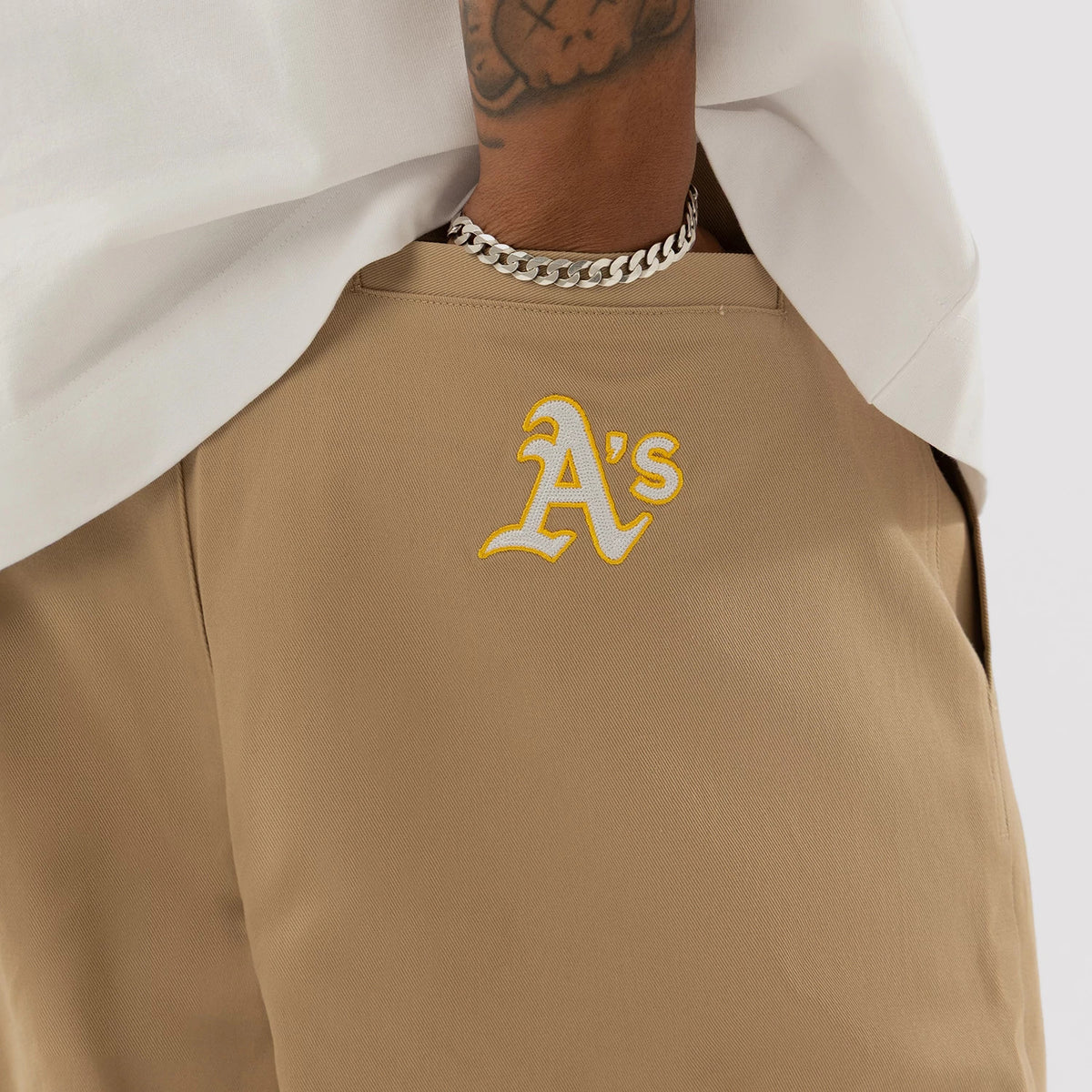 New Era Athletics MLB Beige Woven Chinos, rear view on a model with a white T-shirt, featuring a yellow and white embroidered Athletics logo on the right back pocket and a prominent chain bracelet on the model’s wrist.