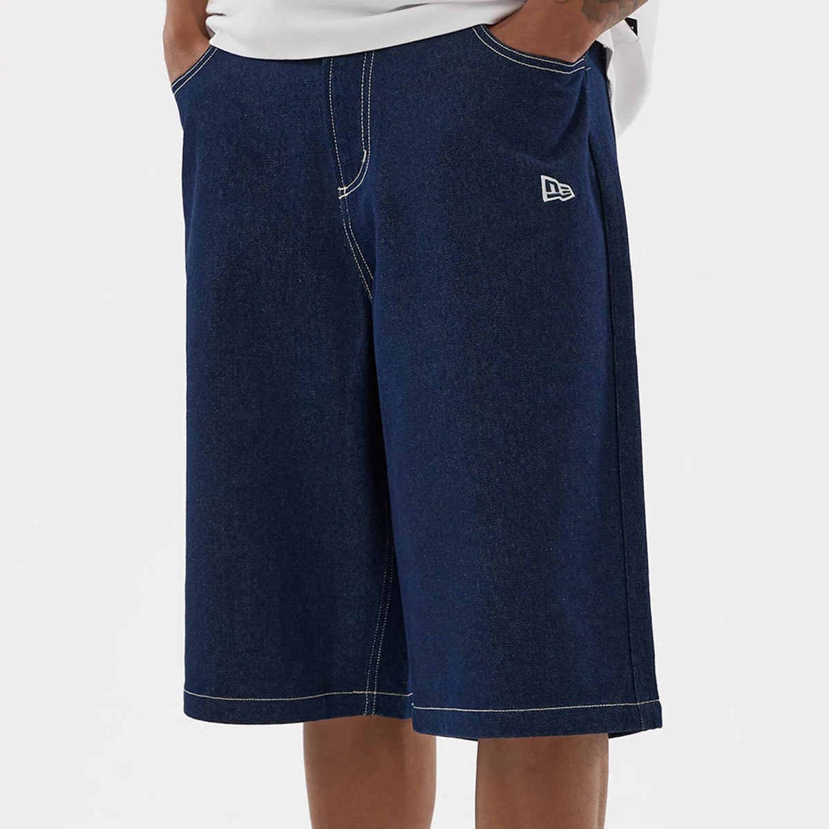 New Era LA Dodgers MLB Jort Dark Blue Oversized Shorts, front view showing dark blue denim with white stitching along the seams and pockets, an embroidered New Era logo on the left side, worn by a model with hands in pockets.