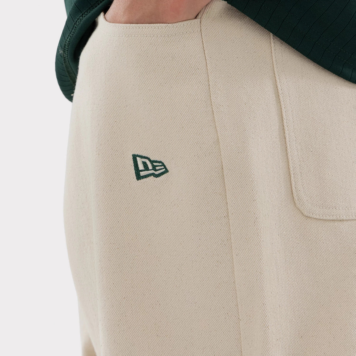 New Era Athletics MLB Jort Cream Oversized Shorts, close-up three-quarter view showing the cream fabric with an embroidered green New Era logo on the side, a large pocket detail, and the bottom of a dark green top overlapping the waistband.