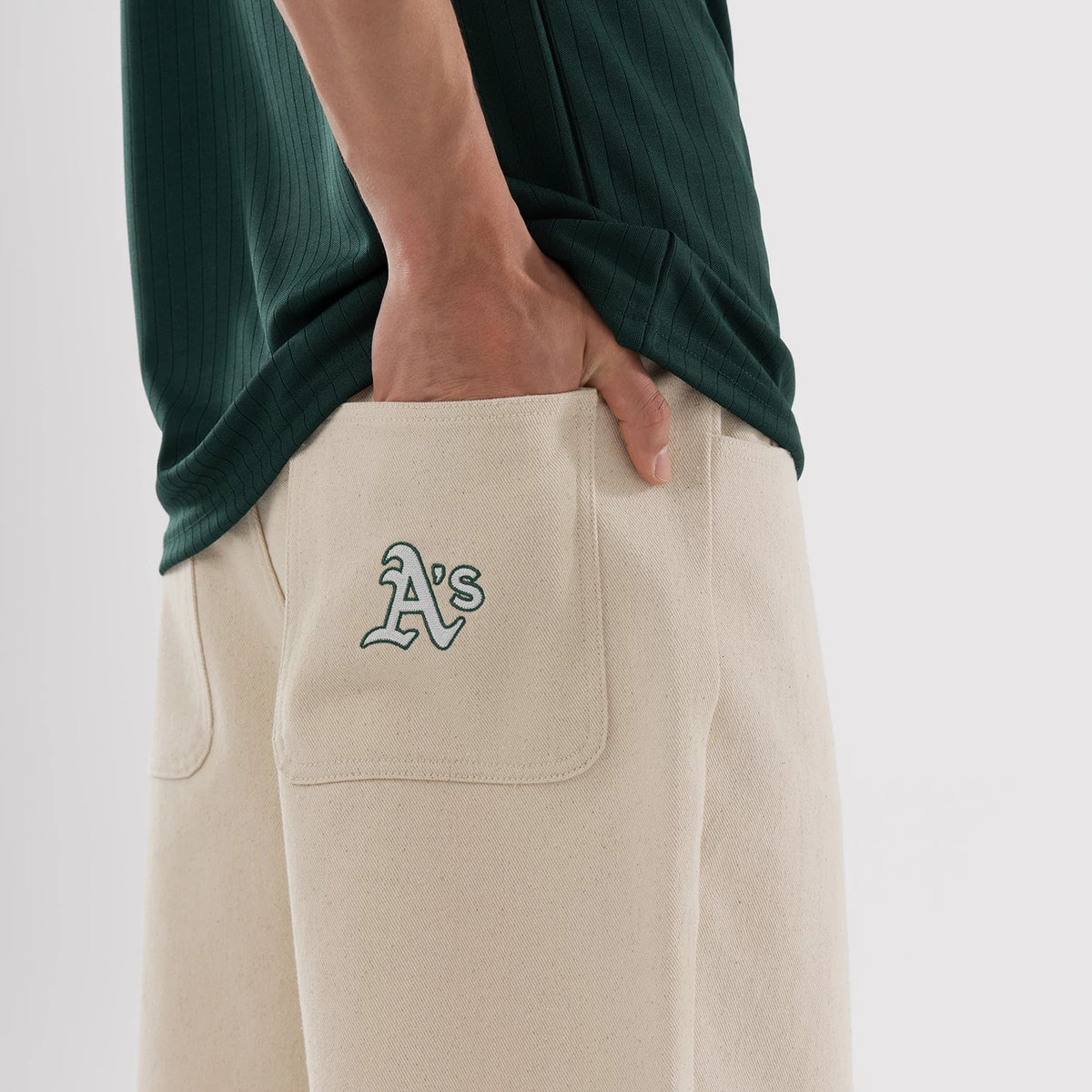New Era Athletics MLB Jort Cream Oversized Shorts, side view showcasing the relaxed fit with a hand in the pocket, cream-coloured fabric, and the green embroidered A's logo on the flap of the back pocket, paired with a dark green top.