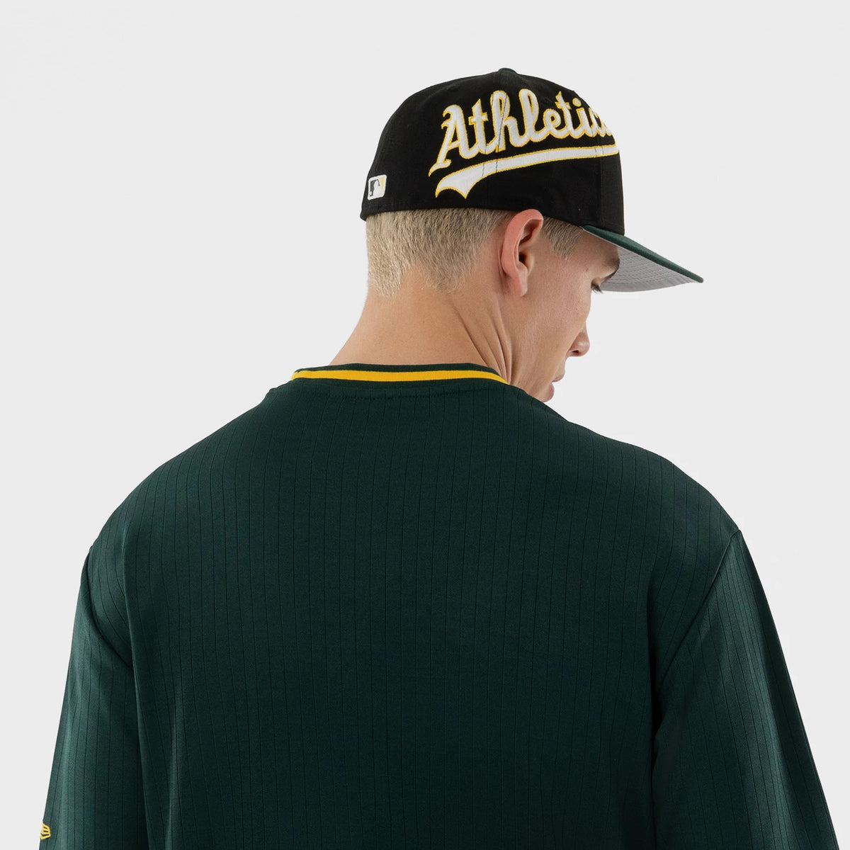 New Era Athletics MLB Dark Green Oversized T-Shirt, back view on a model with a black cap featuring oversized embroidered Athletics lettering; the dark green fabric has vertical lines and a contrasting yellow trim at the back neckline and sleeves.
