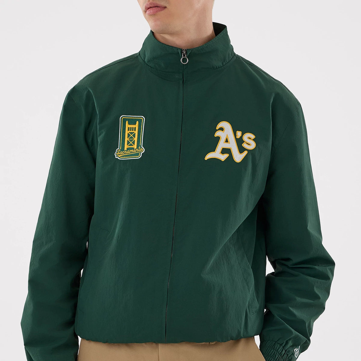 New Era Athletics MLB Dark Green Sports Jacket, front view on a model showing the zippered front, an embroidered A's logo on the left chest, and a decorative patch with Sacramento lettering on the right chest.