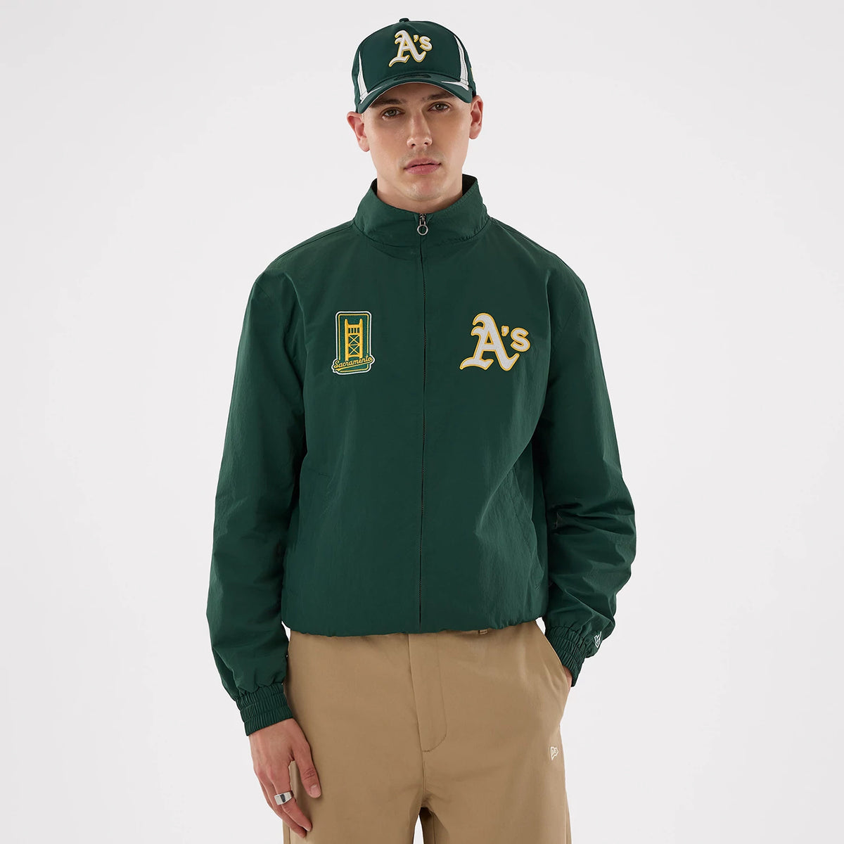 New Era Athletics MLB Dark Green Sports Jacket, front view on-model showcasing a deep green zip-up jacket with embroidered A's logo on the left chest, an additional patch on the right, and a matching cap worn with beige trousers.