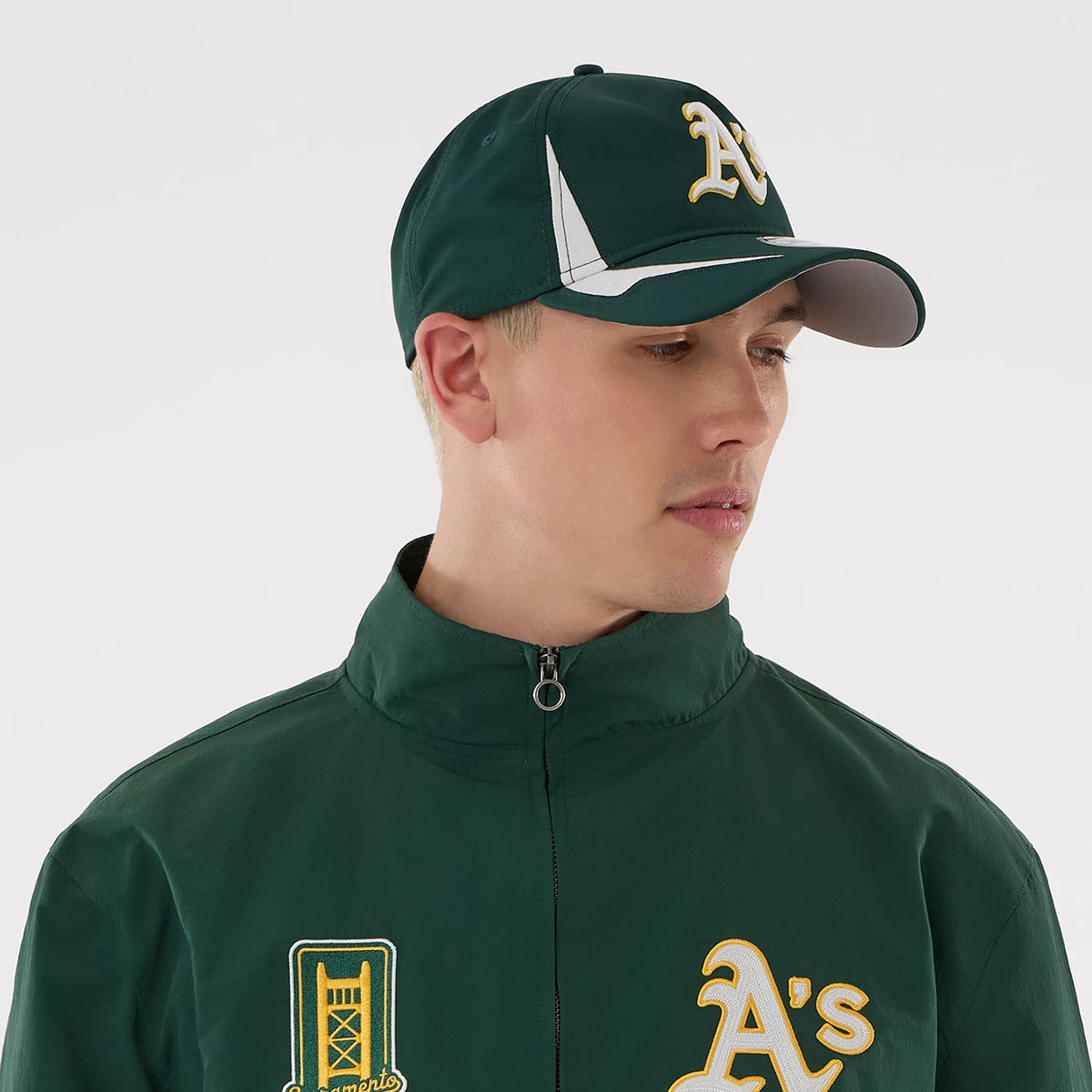 New Era Athletics MLB Dark Green Sports Jacket, front three-quarter view on a model also wearing a dark green cap, showing the zippered jacket with yellow embroidered A's logo on one side and a tower design embroidered on the other side.