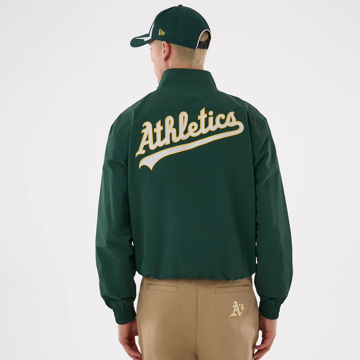New Era Athletics MLB Dark Green Sports Jacket, back view on a model showing the large embroidered Athletics script in cream and gold across the shoulders, paired with tan trousers featuring an embroidered A's logo above the right pocket.