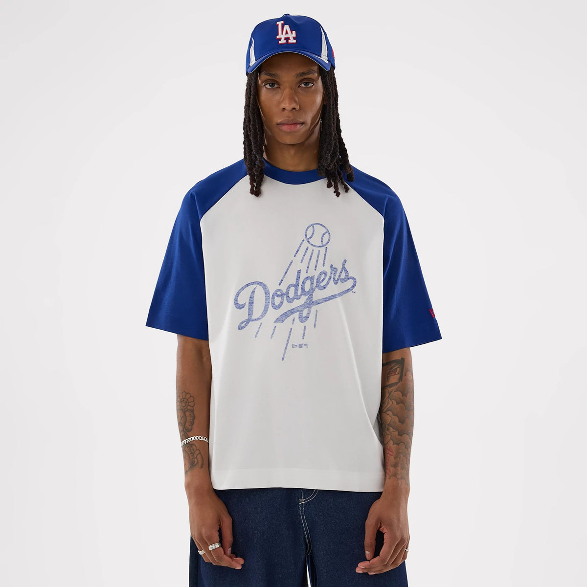 New Era LA Dodgers MLB White Boxy T-Shirt, front view on a model featuring a blue Dodgers graphic with a baseball and lines suggesting motion, blue raglan sleeves, and a small red embroidered logo on the left sleeve, paired with a blue cap.