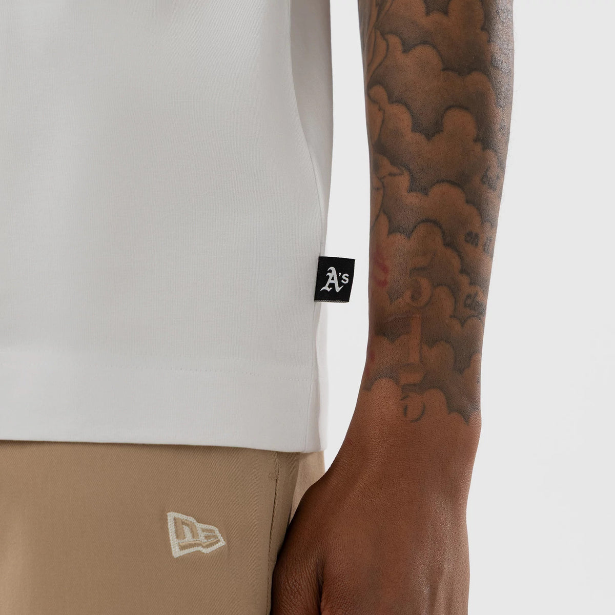 New Era Athletics MLB White Boxy T-Shirt, close-up view showing a black label with the A's logo on the lower side hem in contrast to beige trousers featuring an embroidered New Era logo, with a tattooed arm next to the garment.