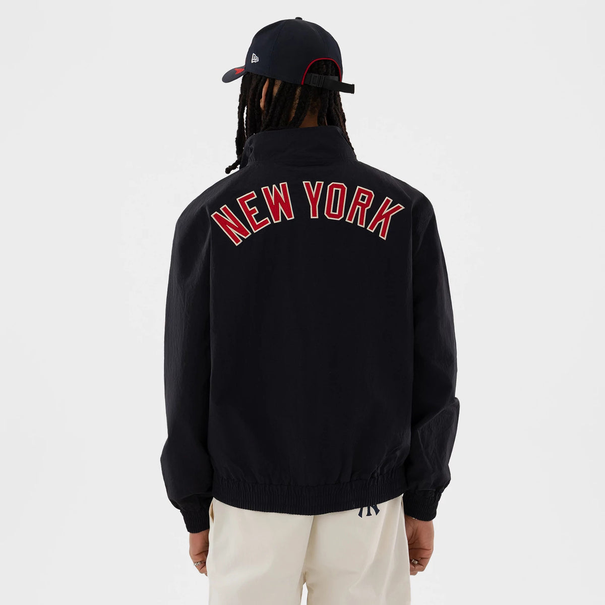New Era New York Yankees MLB Navy Sports Jacket, back view on a model wearing a matching navy cap and light trousers, featuring prominent red embroidered NEW YORK lettering across the shoulders of the navy jacket and a gently elasticated hem.