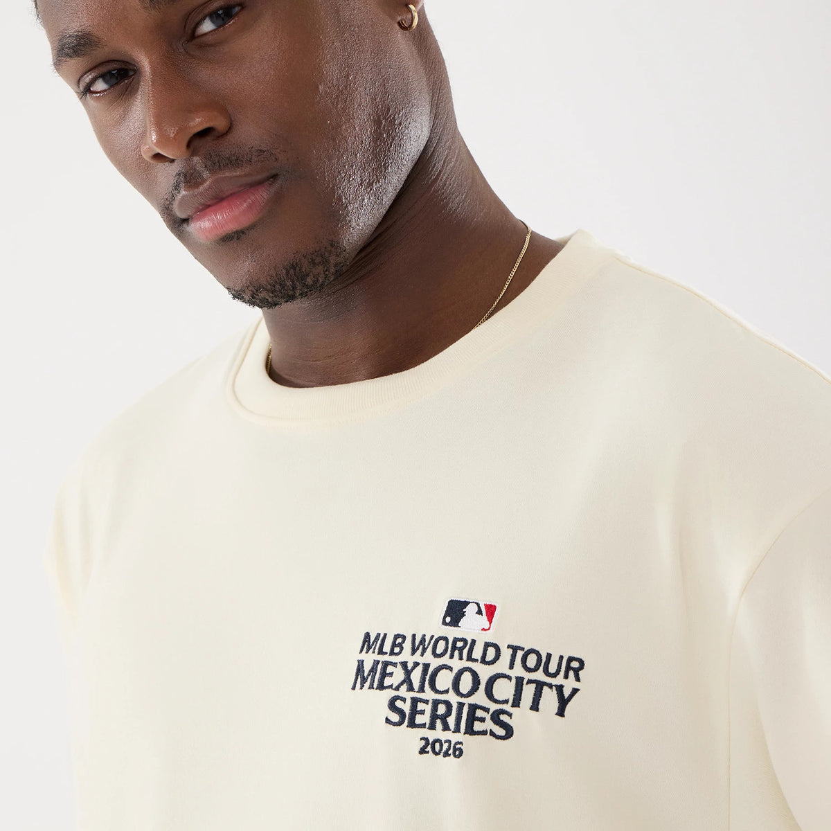 New Era Dueling Logo MLB Mexico City Series 2026 Script Cream T-Shirt, three-quarter view on-model showing a close-up of the chest with an embroidered MLB World Tour Mexico City Series 2026 logo in dark lettering next to a small coloured MLB emblem.