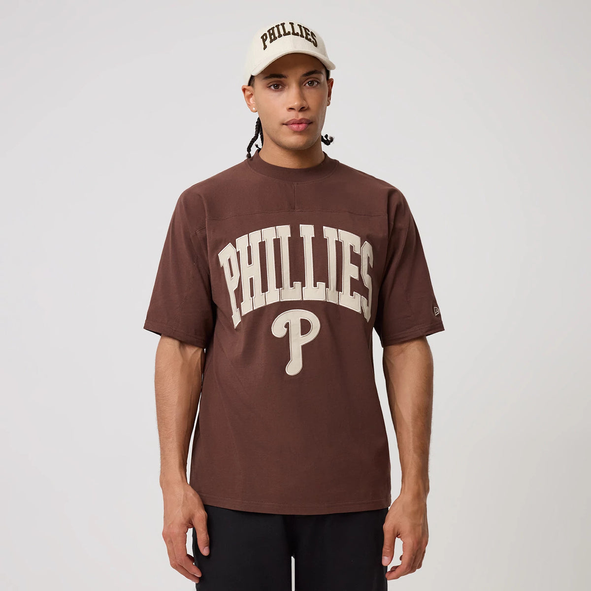 New Era Philadelphia Phillies MLB Arch Graphic Brown Oversized T-Shirt, front view on a model styled with a matching cap, featuring large printed PHILLIES lettering arching across the chest and a raised Phillies P logo beneath.