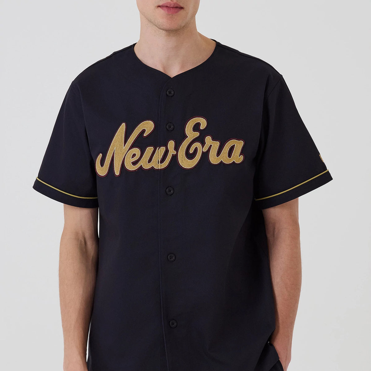 New Era Navy Straight Hem Jersey, front view on a model featuring large gold embroidered New Era lettering across the chest, black button closures down the centre, and gold trim around the short sleeves.