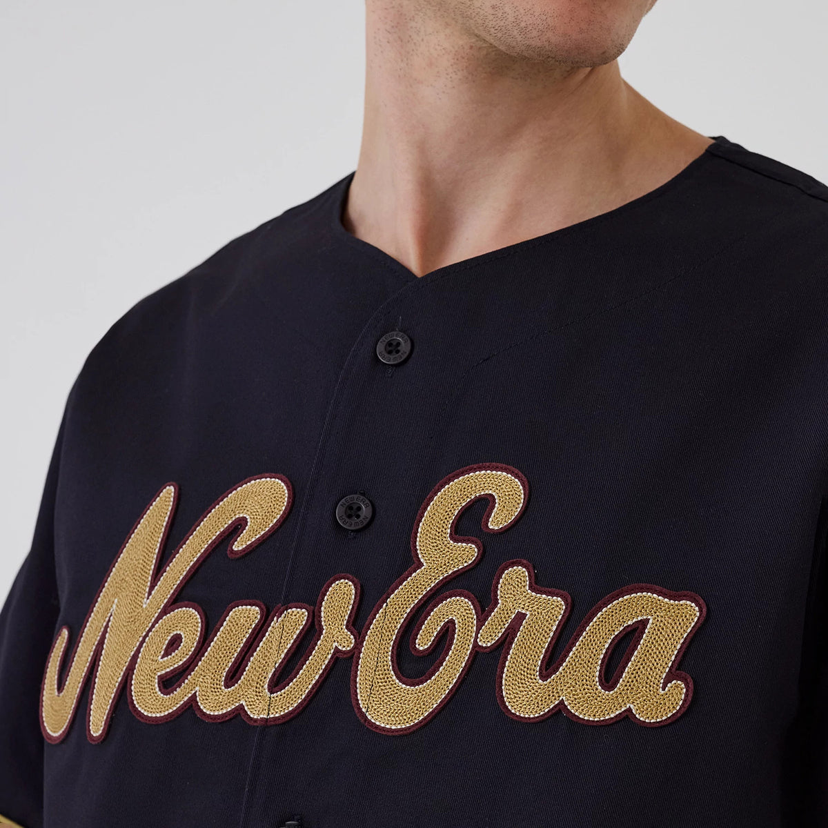 New Era Navy Straight Hem Jersey, front view on a model showing large gold embroidered New Era script across the chest, with contrasting burgundy outline, and black button detail along the centre front of the navy jersey.