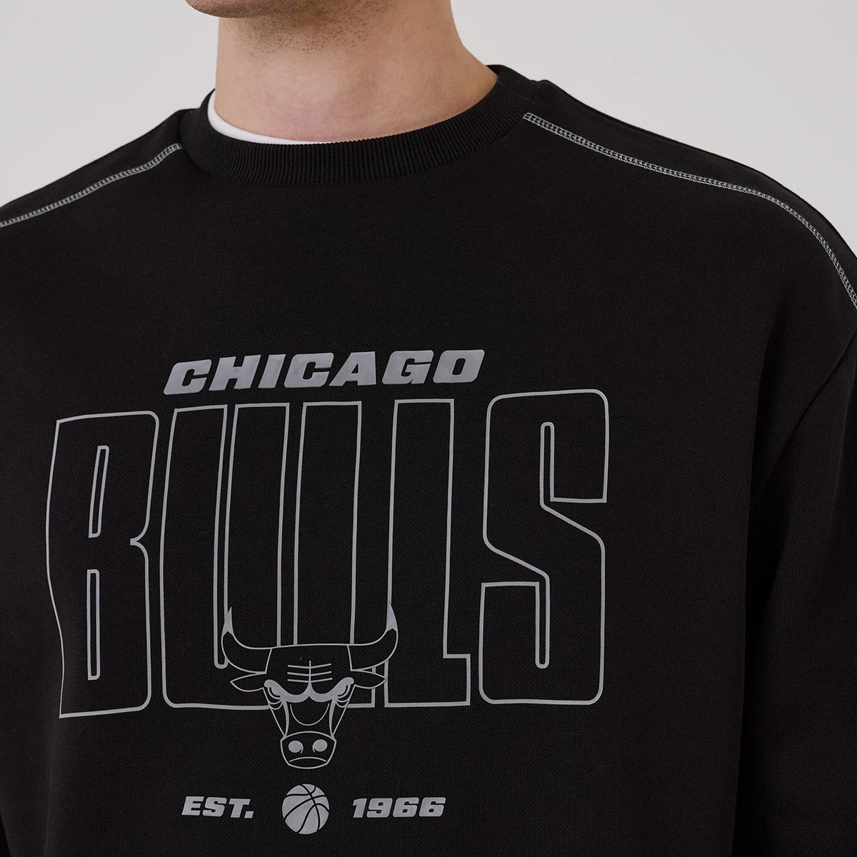 New Era Chicago Bulls NBA Graphic Black Crew Neck Sweater, front view on a model featuring large grey Chicago Bulls text and a bull head logo across the chest, with contrasting stitch details on the shoulders and a ribbed neck.