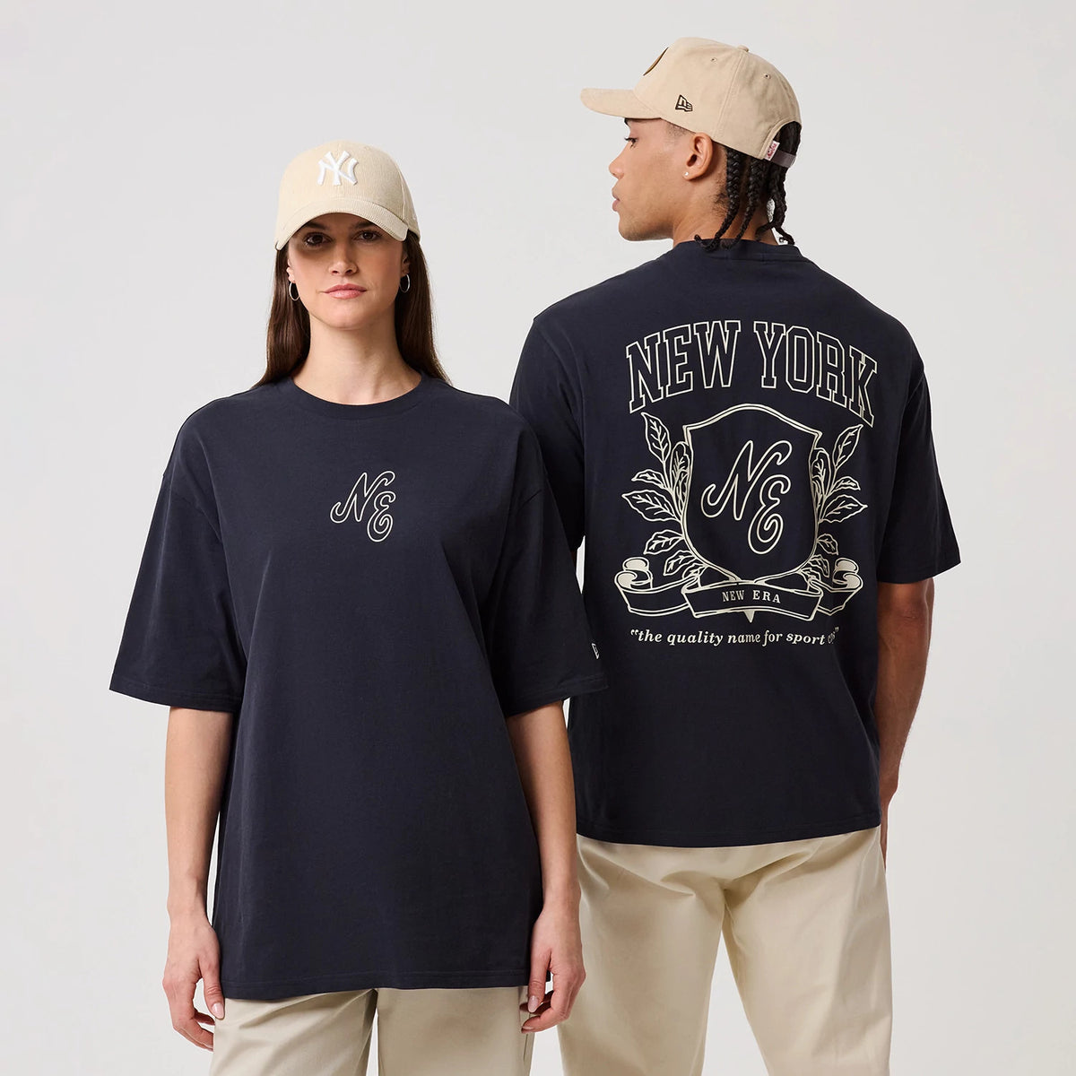 New Era Crest Graphic Navy Oversized T-Shirt, on-model view showing two models in beige caps, one facing forward with an NE printed logo on the chest and the other with a detailed New York crest graphic and lettering on the back.