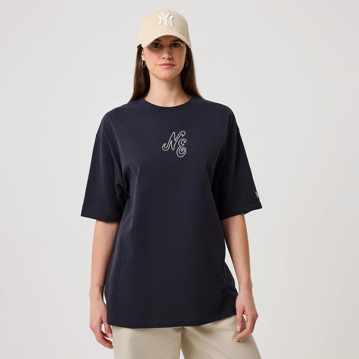 New Era Crest Graphic Navy Oversized T-Shirt, front view on a model with cream trousers and a beige cap, showing an embroidered NE crest on the chest and a small embroidered New Era logo on the left sleeve above the hem.