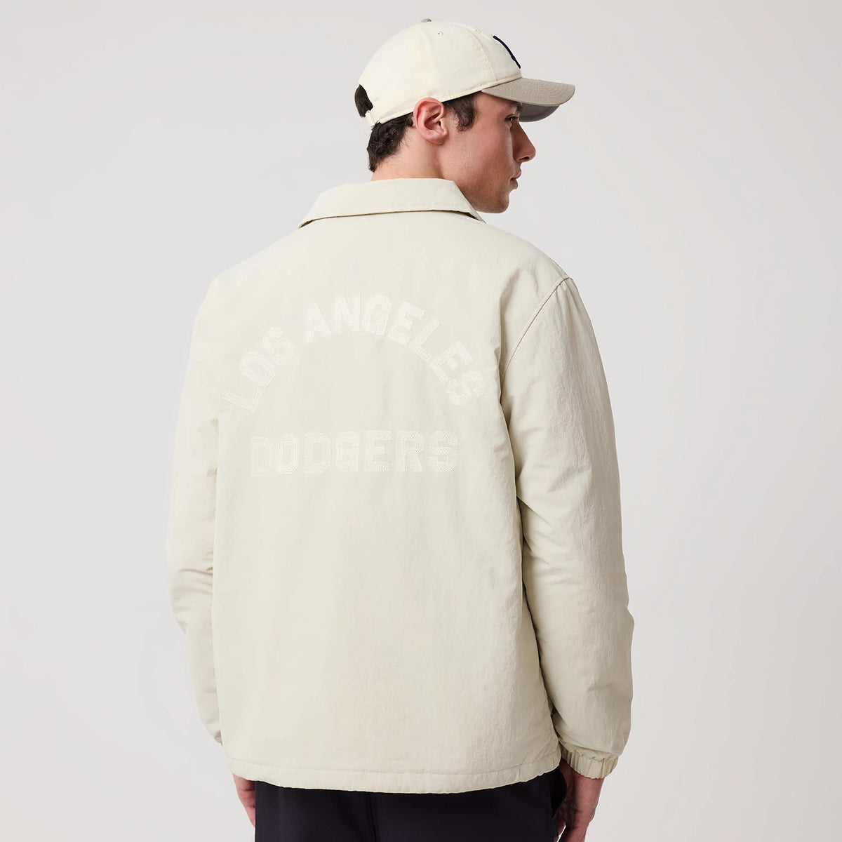 New Era LA Dodgers MLB Light Beige Coach Jacket, back view on a model wearing a matching cap, showing light beige fabric with embroidered Los Angeles Dodgers in an arch shape across the upper back, set against simple, clean jacket lines.