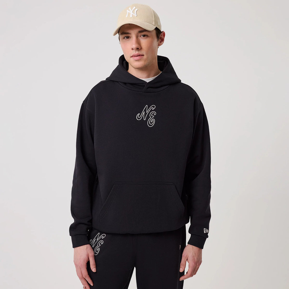 New Era Crest Graphic Navy Pullover Hoodie, front view on a model paired with a cream cap, featuring an embroidered NE crest centred on the chest, a spacious front pocket, and a embroidered New Era logo near the left cuff.