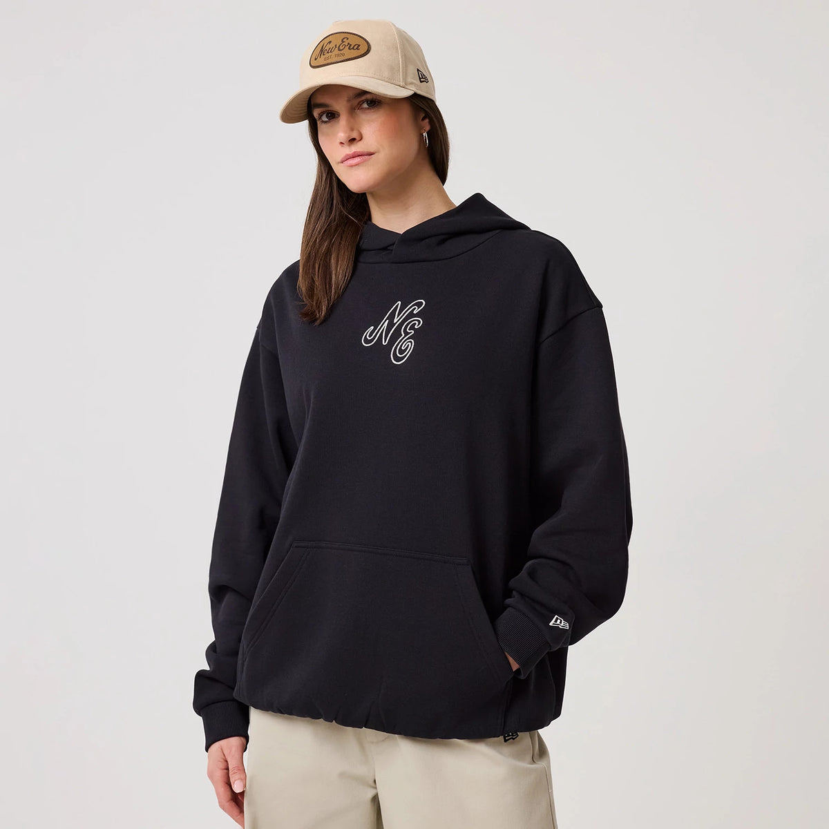 New Era Crest Graphic Navy Pullover Hoodie, front view on-model displaying a navy hoodie with an embroidered NE crest on the chest, a kangaroo pocket, and a small New Era logo on the left sleeve, paired with a beige cap.