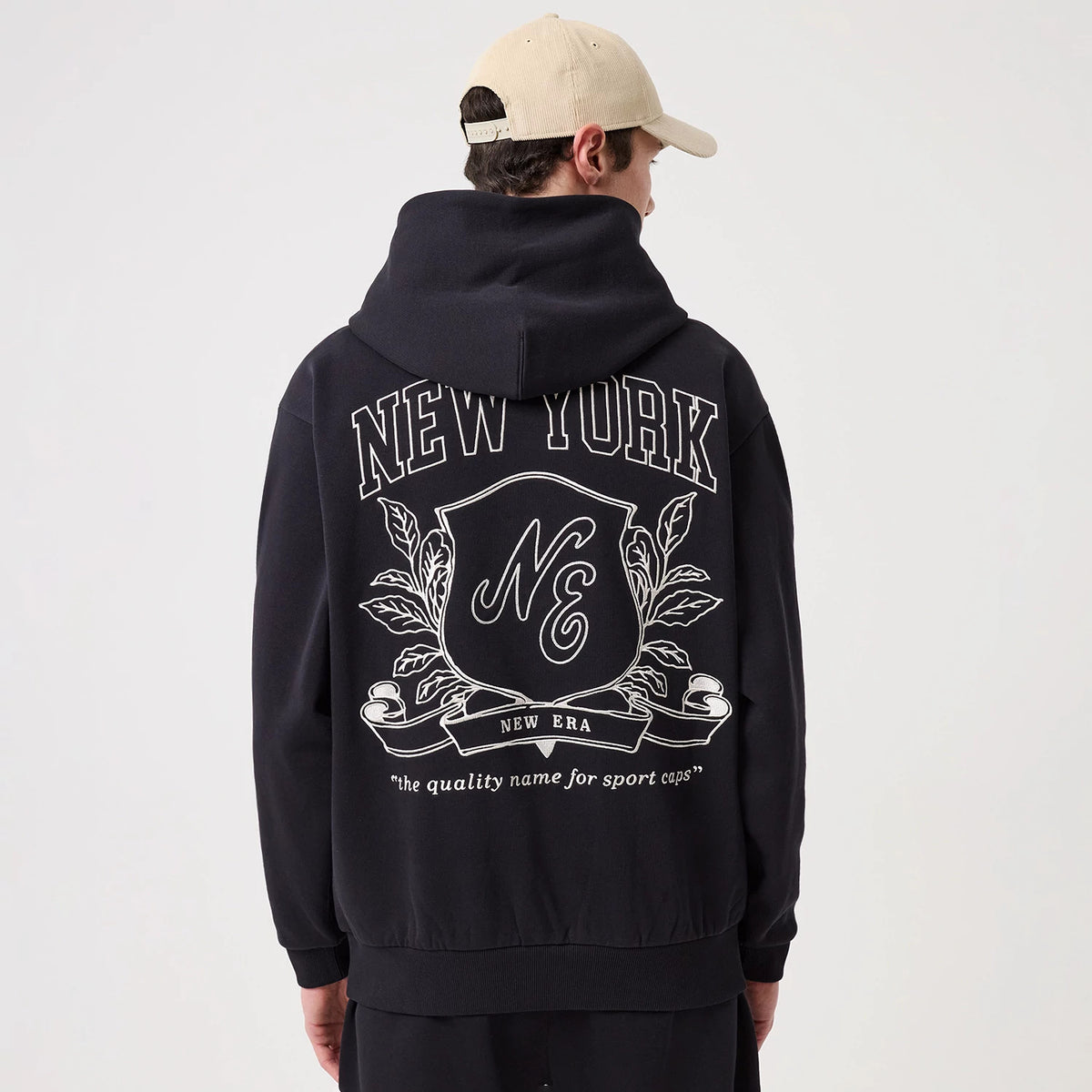 New Era Crest Graphic Navy Pullover Hoodie, back view showing a large crest graphic with NE lettering in the centre, NEW YORK across the shoulders, and New Era text below, worn on a model styled with a beige cap.