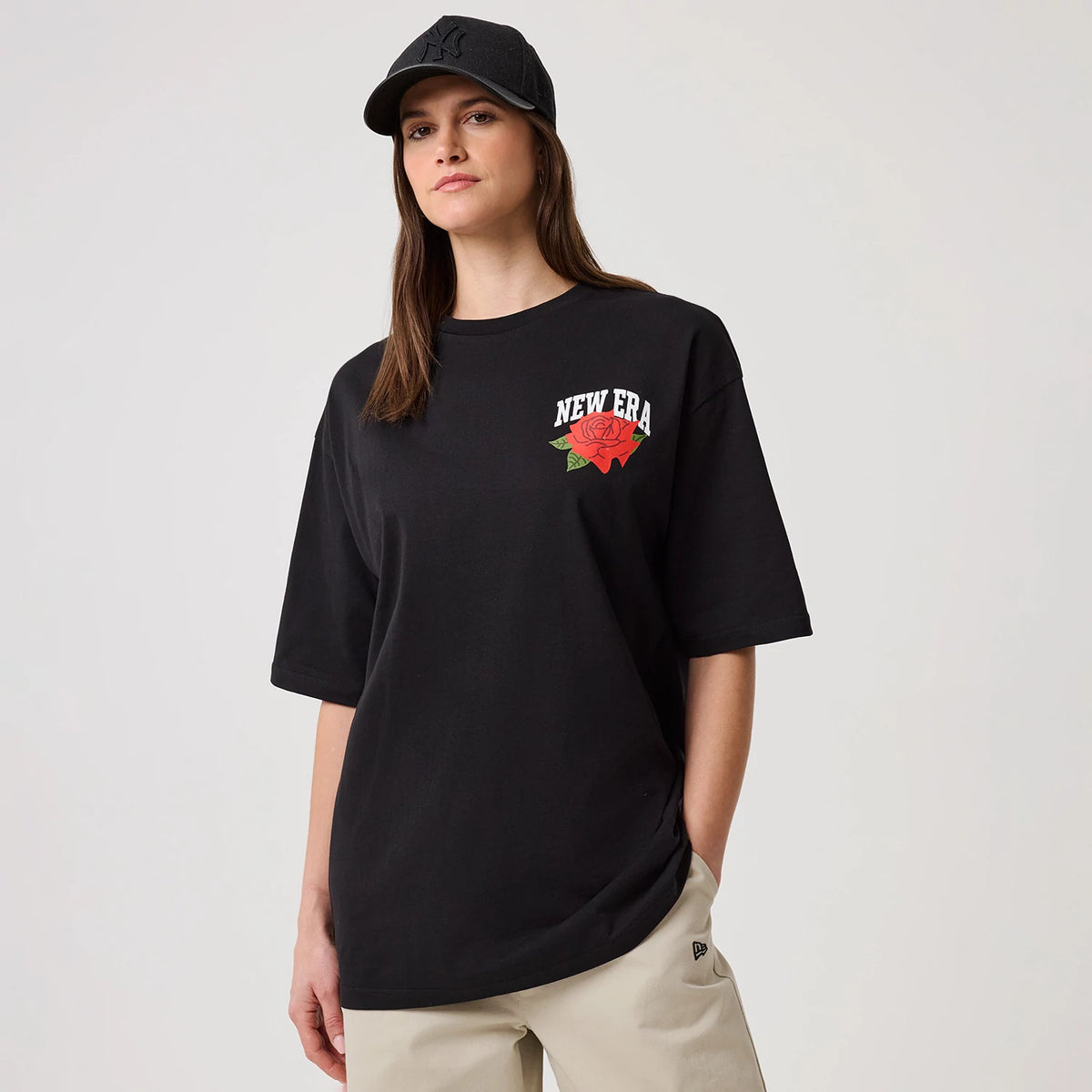 New Era Floral Graphic Black T-Shirt, front view on a model wearing a black cap, featuring a white embroidered New Era logo with a red rose on the left chest and classic short sleeve design paired with neutral trousers.