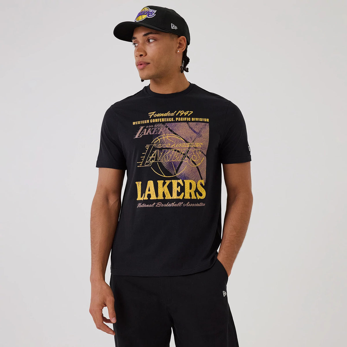 New Era LA Lakers NBA Graphic Black T-Shirt, front view on-model featuring a Lakers graphic centred on the chest with Founded 1947 above it, contrasting yellow logo embroidery on the left sleeve, paired with a matching black cap.