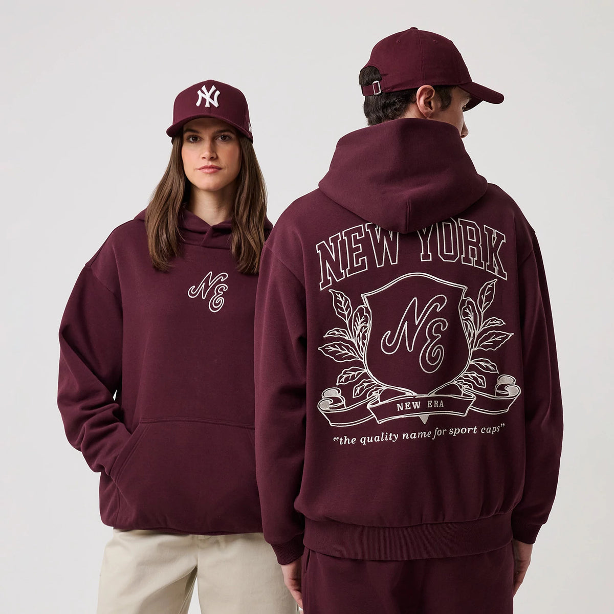 New Era Crest Graphic Dark Purple Pullover Hoodie, front and back view showing the embroidered NE crest logo on the front left chest and a large graphic on the back with New York lettering above a crest, worn with a matching dark purple cap.