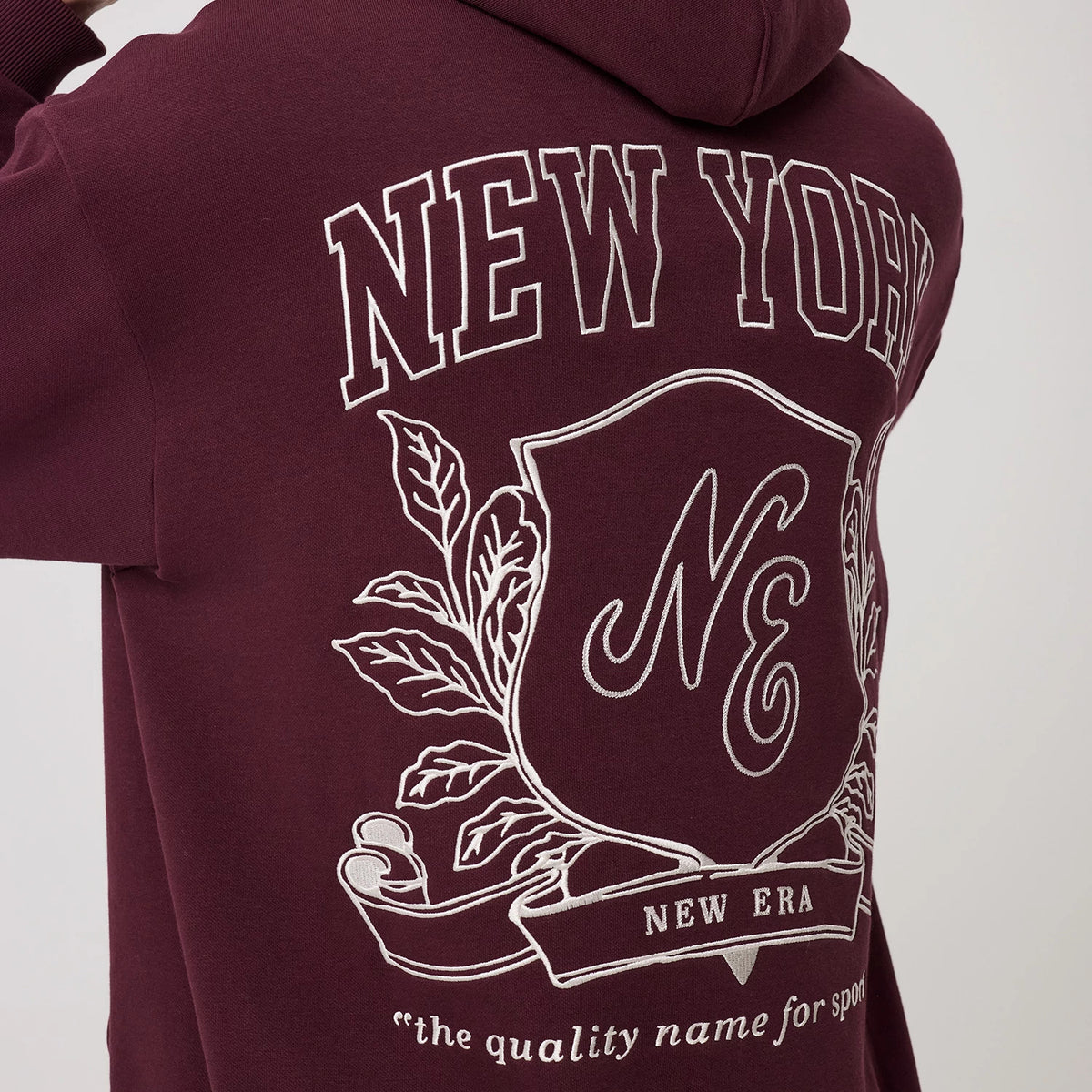 New Era Crest Graphic Dark Purple Pullover Hoodie, back view displaying an embroidered New York crest with decorative leaves and the New Era lettering, above the phrase the quality name for sports at the base of the dark purple hoodie.
