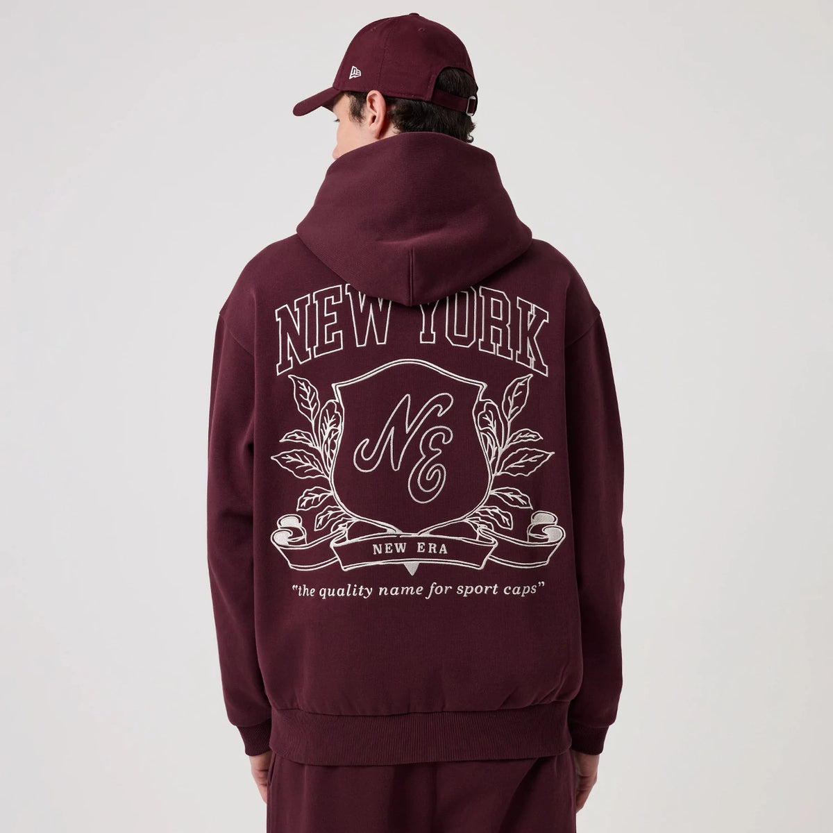 New Era Crest Graphic Dark Purple Pullover Hoodie, back view on a model with a matching dark purple cap, featuring large white New York lettering across the shoulders and an intricate crest graphic with New Era branding and a decorative emblem.