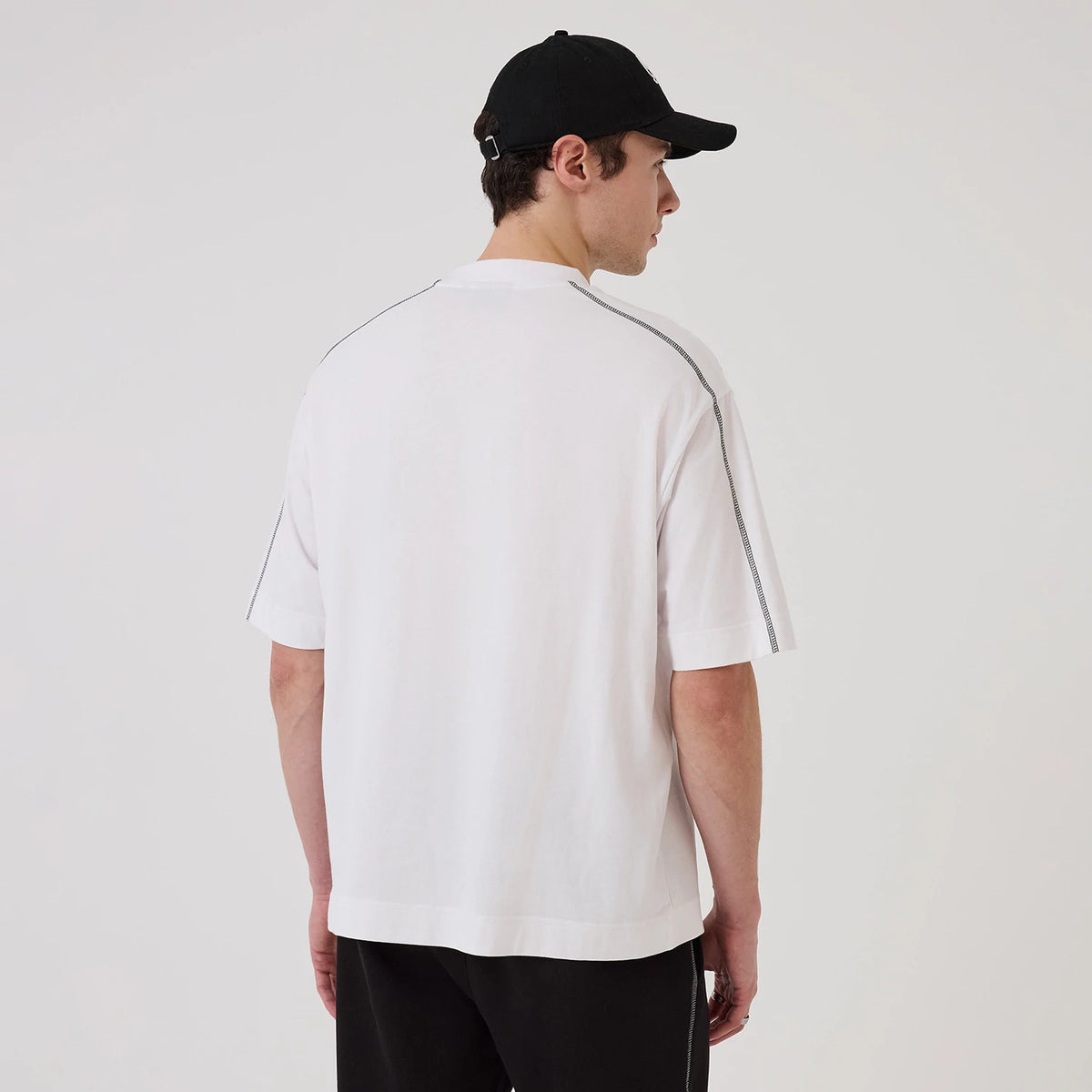 New Era Chicago Bulls NBA Graphic White Boxy T-Shirt, back view on a model showing the plain white back with short sleeves, and piping detail along the shoulder seams, styled casually with black trousers and a black cap.