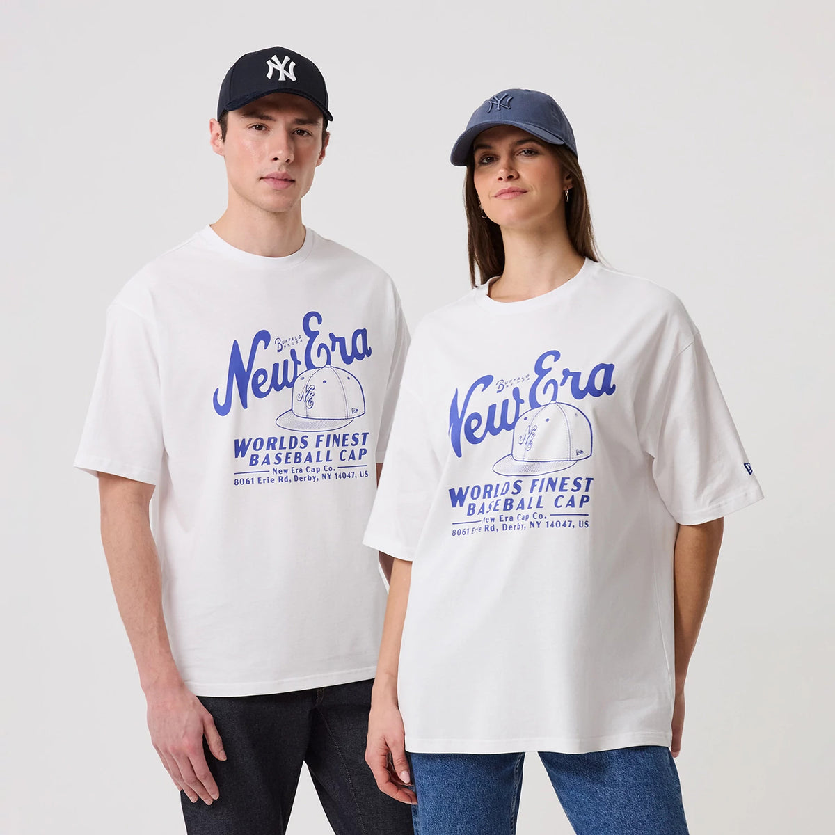 New Era Heritage White Oversized T-Shirt, front view on-model showing the blue New Era graphic across the chest, a baseball cap illustration beneath, and oversized short sleeves; the models also wear caps, adding a casual styling element.