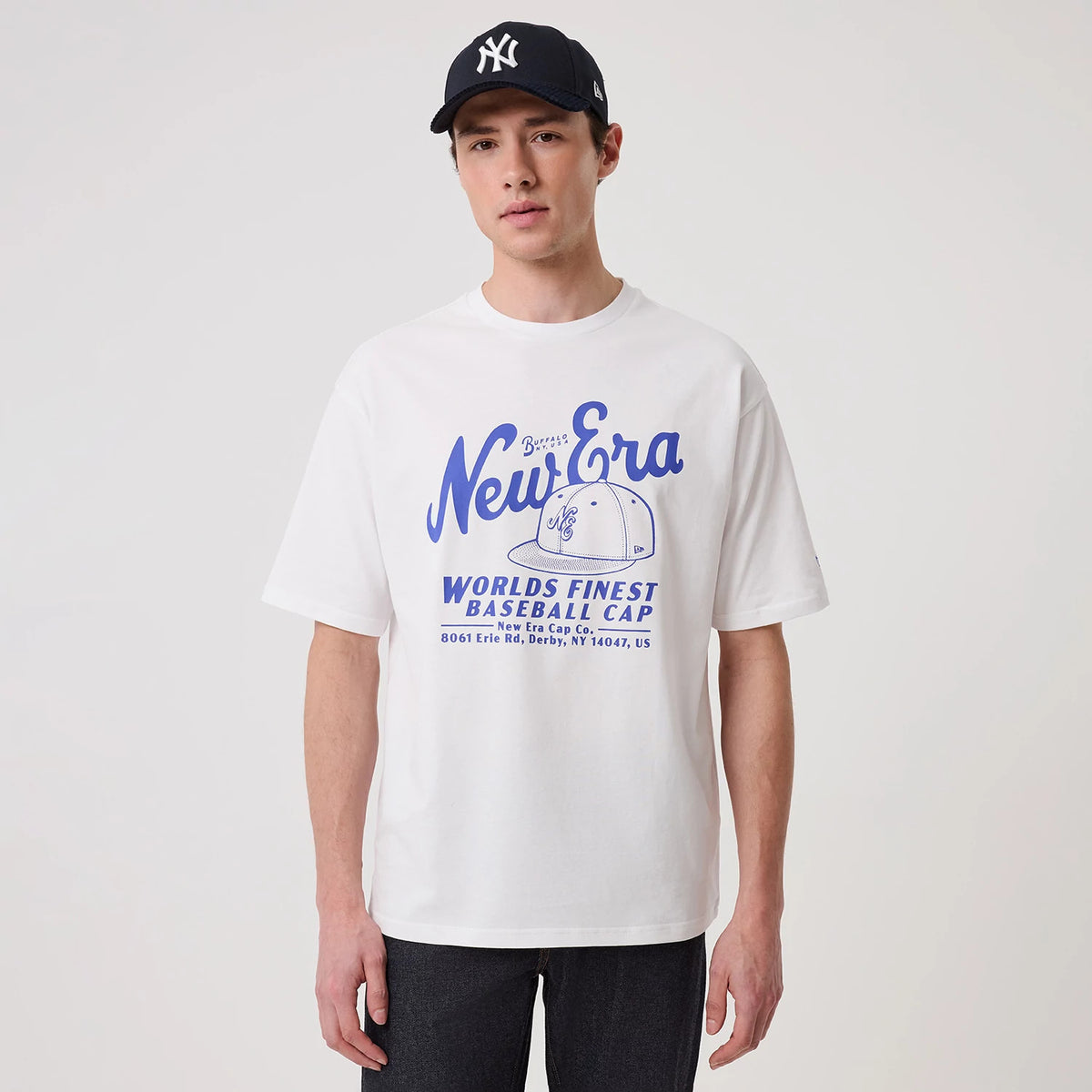 New Era Heritage White Oversized T-Shirt, front view on-model showing a blue printed New Era logo across the chest with a cap graphic and the phrase World's Finest Baseball Cap below, styled with a dark cap and trousers.