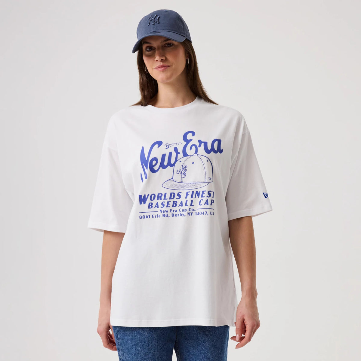 New Era Heritage White Oversized T-Shirt, front view on-model featuring a large blue graphic with New Era branding and a cap illustration on the chest, a small embroidered logo on the left sleeve, paired with a blue cap and jeans.