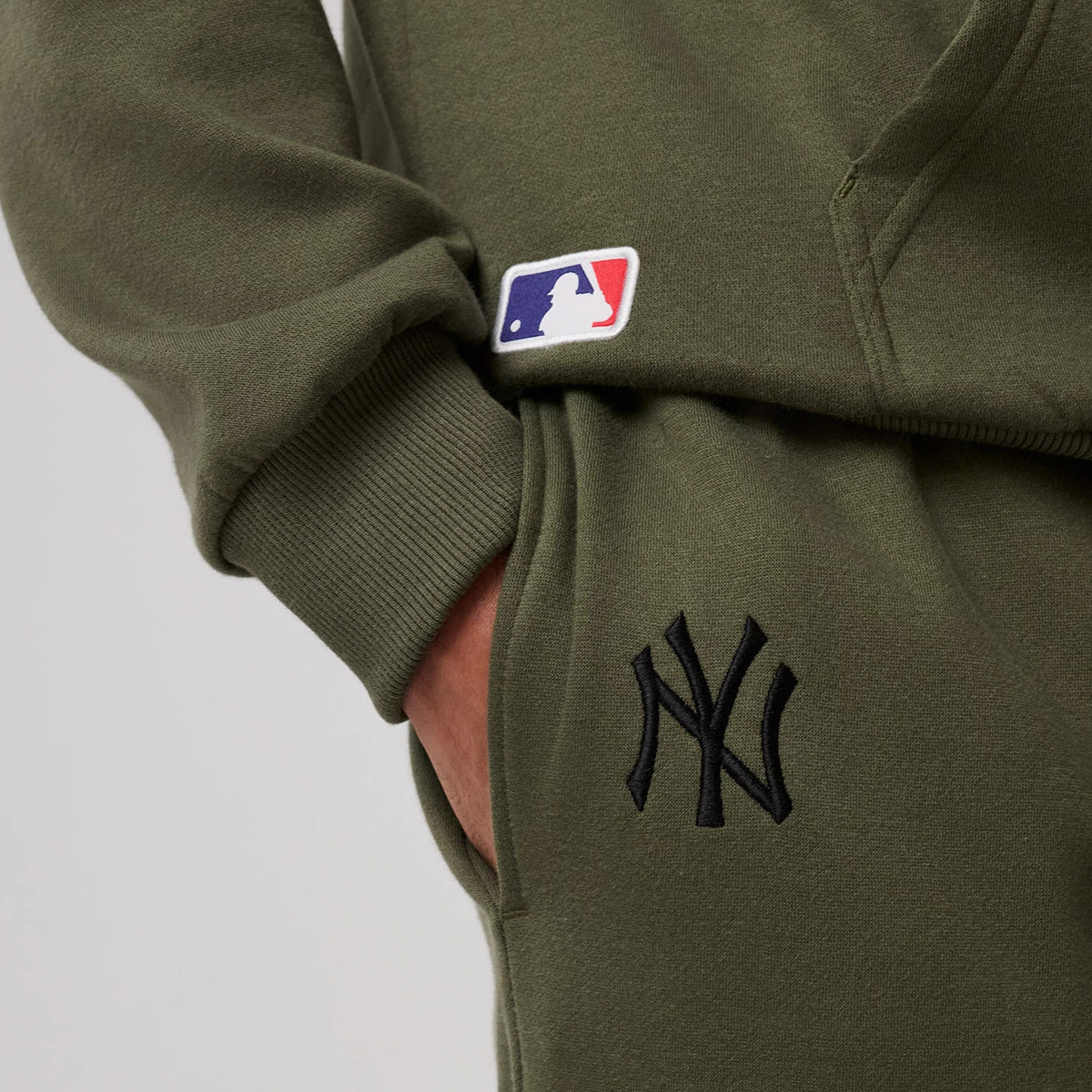 New Era New York Yankees MLB League Essential Green Joggers, close-up showing the black embroidered NY logo on the right thigh, the ribbed cuff around the wrist, and the MLB logo patch near the waistband with a hand tucked in the pocket.