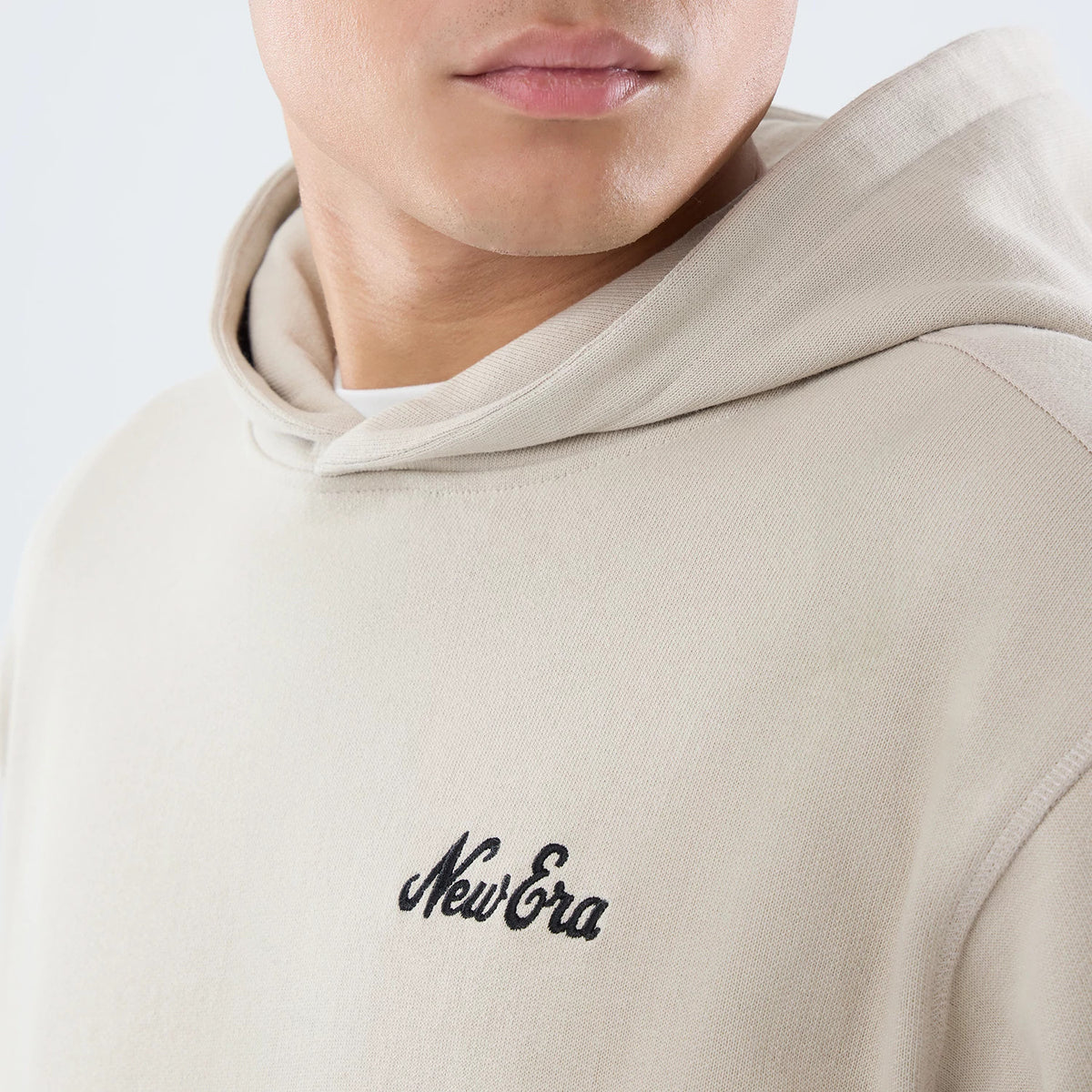 New Era Open Hem Light Beige Pullover Hoodie, front view on a model showing a soft beige colour with a raised black New Era logo on the left chest, and an open hem design near the hood for a relaxed fit.