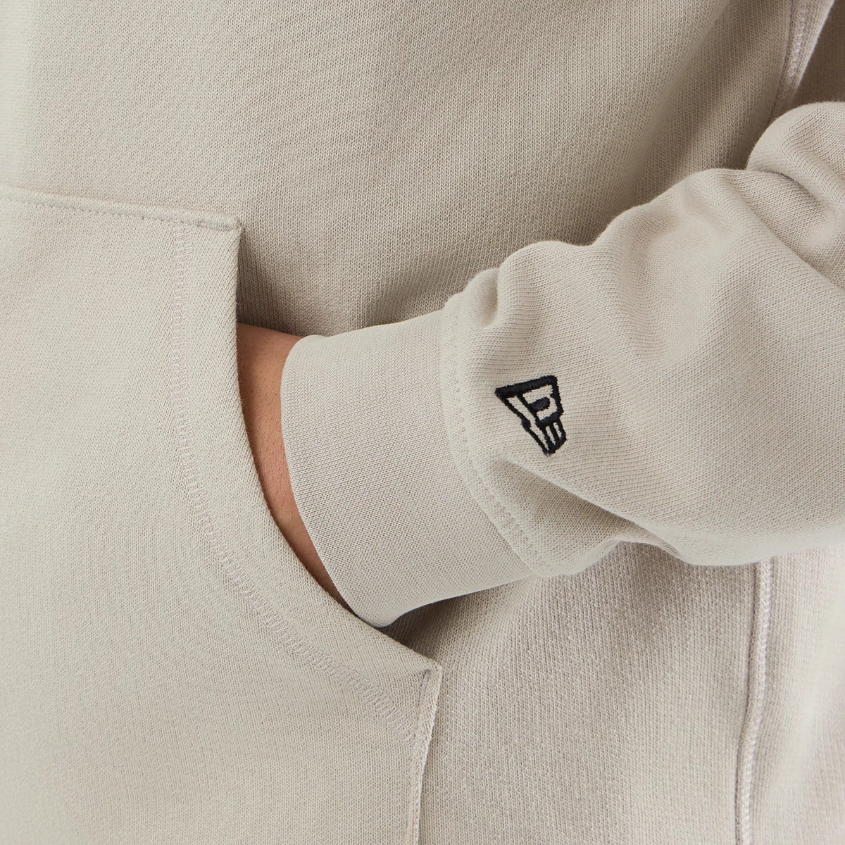 New Era Open Hem Light Beige Pullover Hoodie, side view showing a close-up of the left sleeve with a black embroidered New Era logo near the cuff, the light beige fabric texture, and a partially front pocket with a hand placed inside.