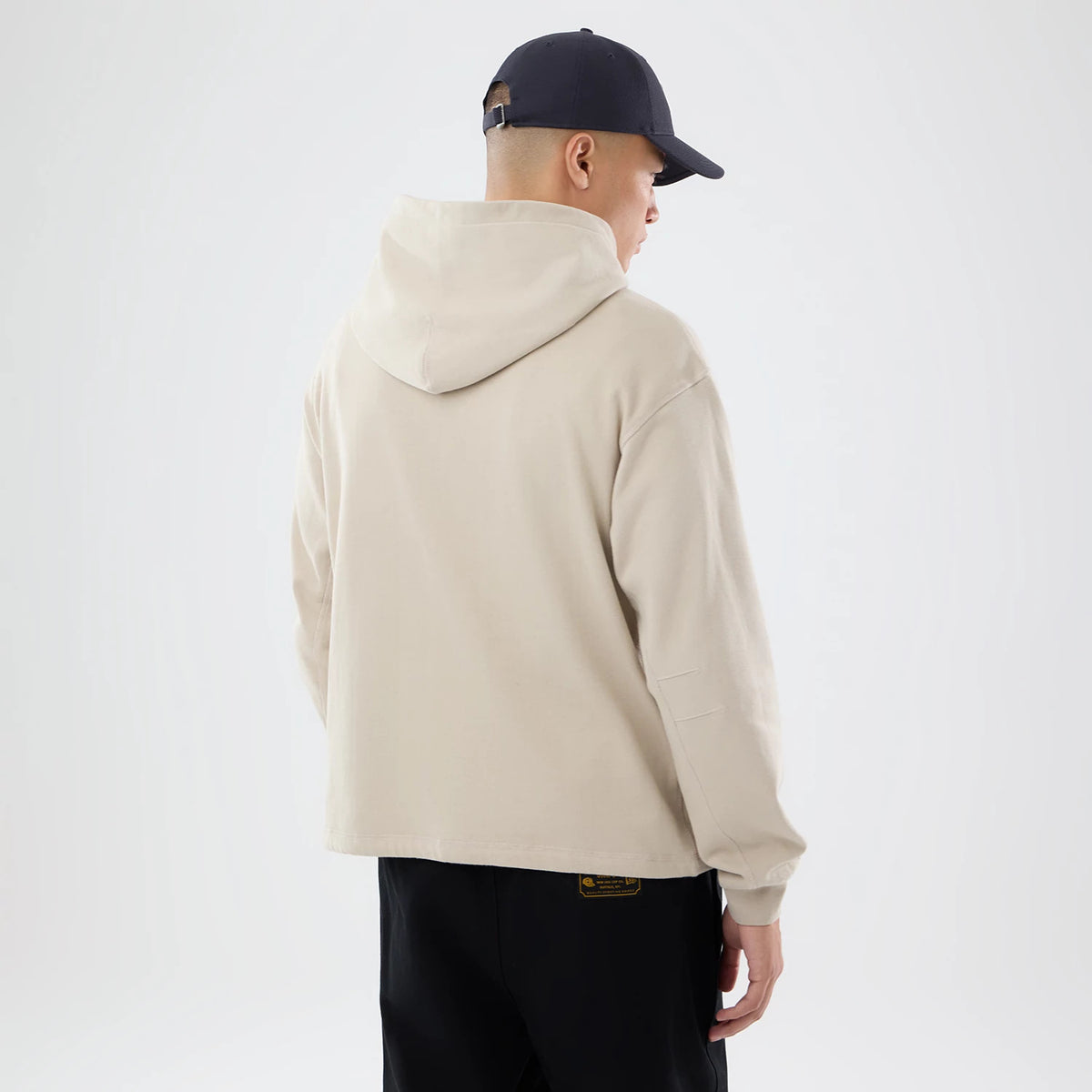 New Era Open Hem Light Beige Pullover Hoodie, back view on a model showing the light beige colour with an open hem and long sleeves, the hood resting naturally on the back, paired with black trousers and a dark cap.