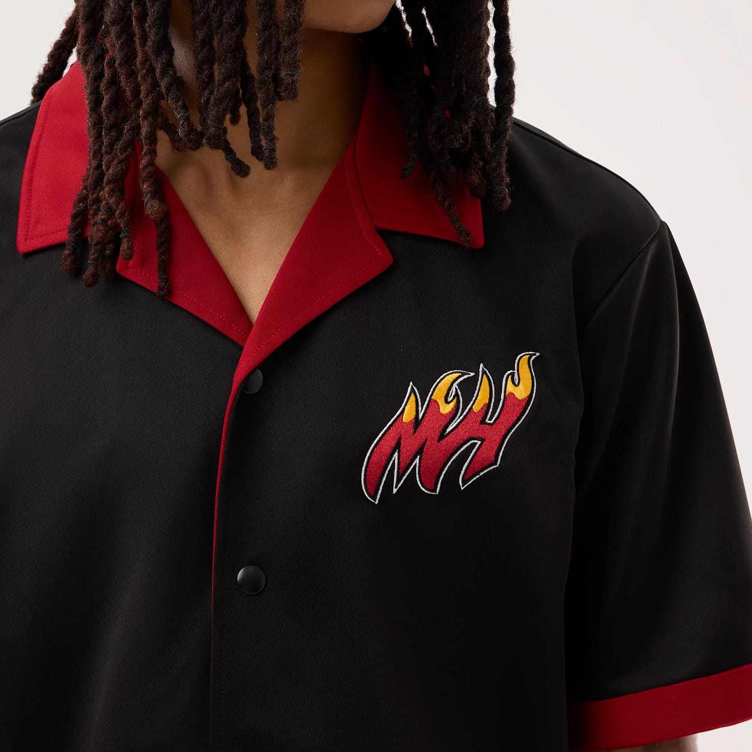 New Era Miami Heat NBA Hardwood Classic Revere Black Short Sleeve Shirt, front view on a model showing the embroidered MH logo with flame details on the left chest, a red collar, and red trim on the short sleeves.