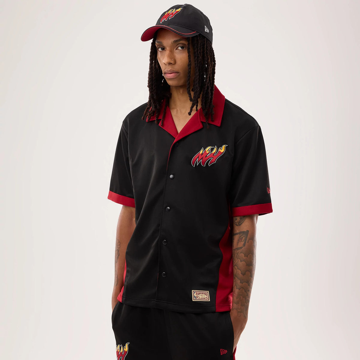 New Era Miami Heat NBA Hardwood Classic Revere Black Short Sleeve Shirt, front view on a model with a matching cap, showing the Miami Heat logo embroidered in flames on the left chest, red accents on the sleeves and collar, and black button closure.