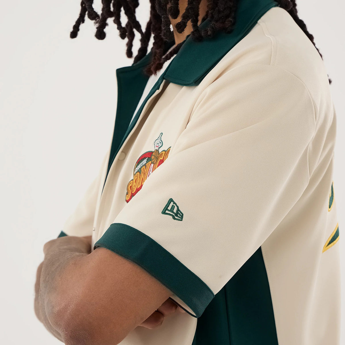 New Era Supersonics NBA Hardwood Classic Revere Beige Short Sleeve Shirt, side view on a model showing the Supersonics logo on the chest, a green New Era flag logo on the left sleeve, and contrasting dark green collar and sleeve trim.